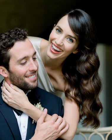 Beautiful bride with Hollywood glam wave hair, natural makeup hugging husband from behind.