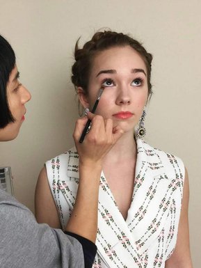 Asian makeup artist using a brush applying eye makeup on actress Holly Taylor, she is wearing a sleeveless top. 