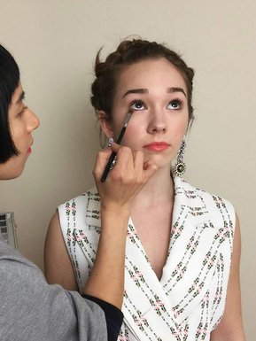 Asian makeup artist using a brush applying eye makeup on actress Holly Taylor, she is wearing a sleeveless top. 