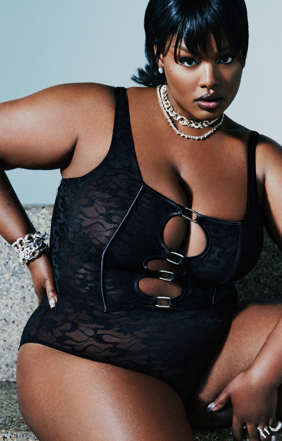American model, Precious Lee, wearing a black corset and underwear, sitting and posing on a stone bench.