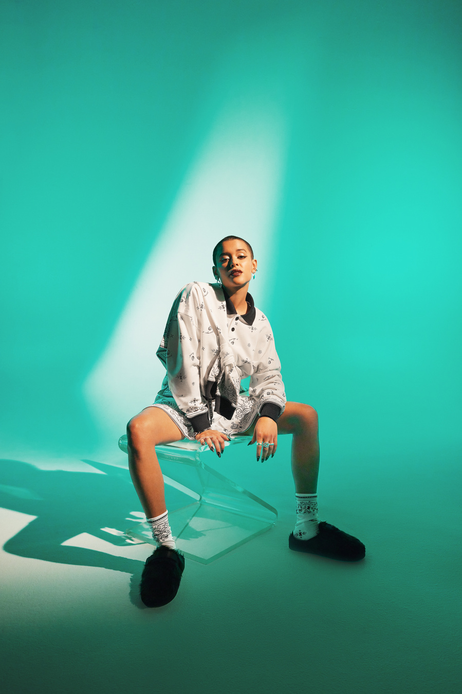 Canadian actress and singer, Jordan Alexander, wearing a white long sleeve, collared shirt and matching long shorts, posing on a chair set against a vibrant teal backdrop