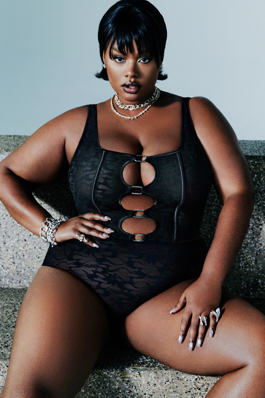 American model, Precious Lee, wearing a black corset and underwear, sitting and posing on a stone bench.