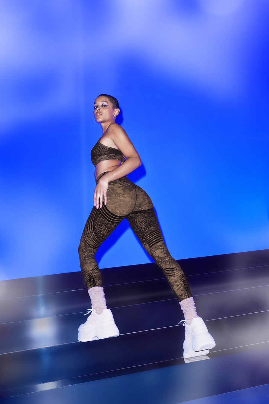 Canadian actress and singer, Jordan Alexander, wearing a tan, lace patterned athletic bra and leggings, posing on an set of stairs against a vibrant blue backdrop
