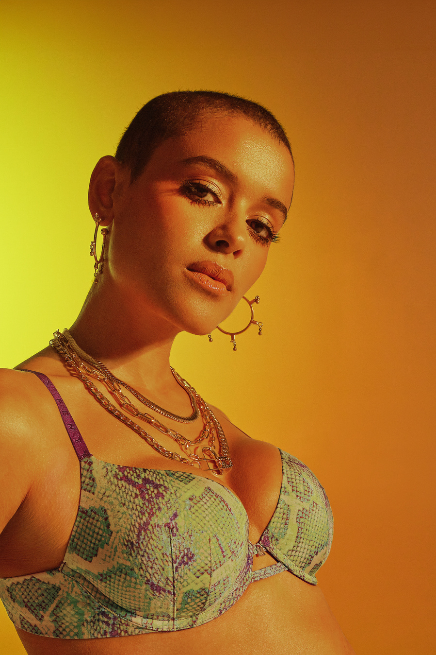 Canadian actress and singer, Jordan Alexander, wearing a green, snake skin patterned bra, posing on an orange and yellow backdrop