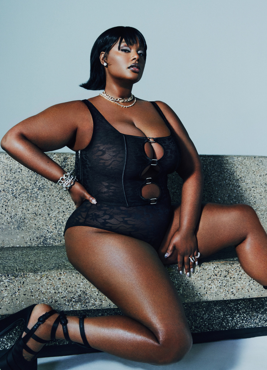 American model, Precious Lee, wearing a black corset and underwear, sitting and posing on a stone bench.