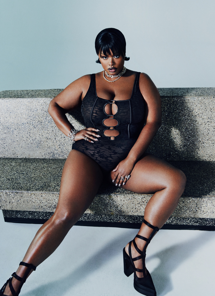 American model, Precious Lee, wearing a black corset and underwear, sitting and posing on a stone bench.