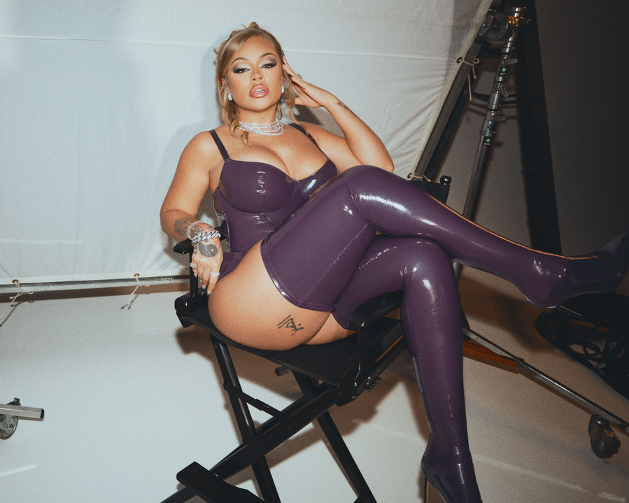 American rapper and celebrity, Latto, wearing purple corset, underwear, and stockings, posing in a studio.