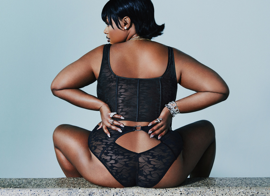 A back shot of the American model, Precious Lee, wearing a black corset and underwear, sitting and posing on a stone bench.