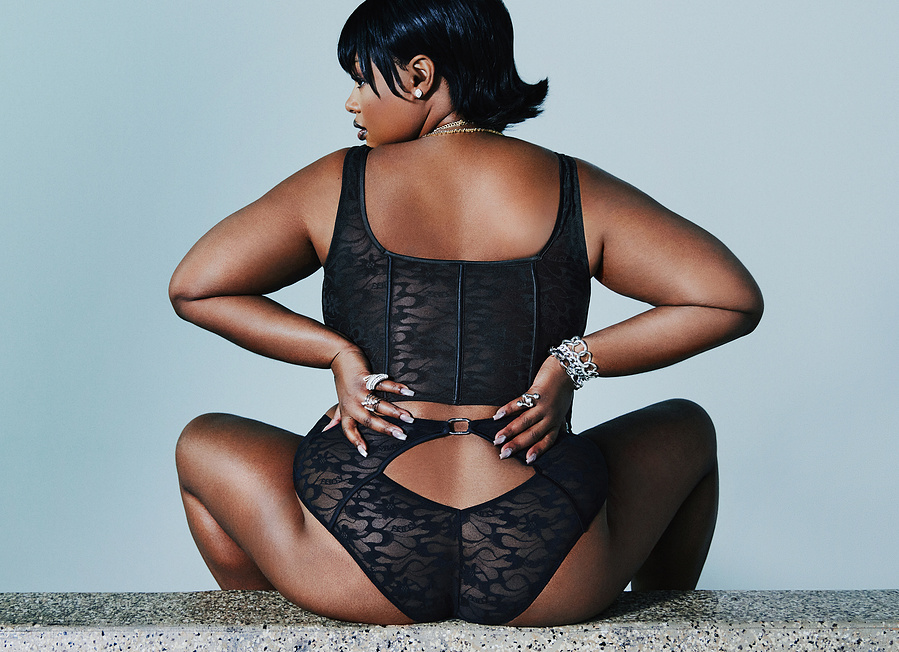 A back shot of the American model, Precious Lee, wearing a black corset and underwear, sitting and posing on a stone bench.