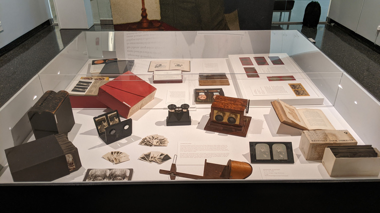 Photo of glass table case with various stereoscopes and accompanying objects. Four kinds of stereoscopes are on display: one at the very front and three in the middle of the case. Own picture taken in the gallery.