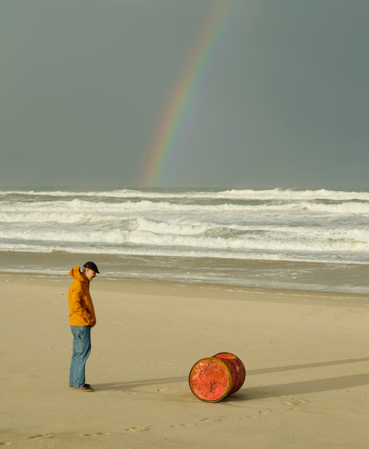 Man on beach staring at an orange oil drum that has been swept ashore with a rainbow in the background