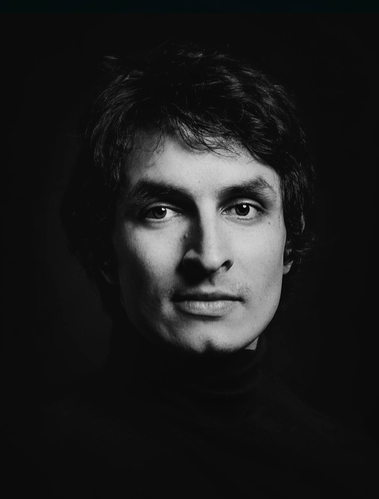 Photo of Paolo Almario's face in grayscale coming out of a dark shaded background.  He has a slight smile and is looking right at the camera.