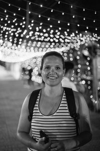 A photo of a woman looking into the lens smiling while sweating, in Vietnam, with blurred lights in the background and the image is black and white.
