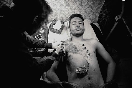 tattoo artist tattooing man, with the man looking straight into the camera, and a dragon tattoo on his chest.