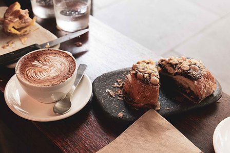 Picture of a crisp chocolate croissant straight out of the oven sitting on black and white stone plate and a latte right next to it featuring latte art.