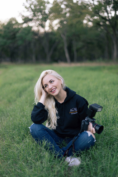 Blonde female photographer sitting on the grass with a camera in hand, smiling and looking away — relaxed outdoor portrait in natural light, Acerra Studio branding visible on hoodie.