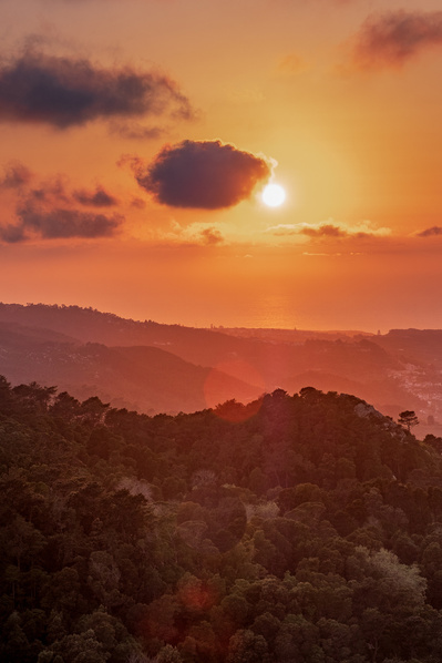 An orange sunset shines above the green, layered hillsides of Sintra, Portugal with a hint of lens flare.