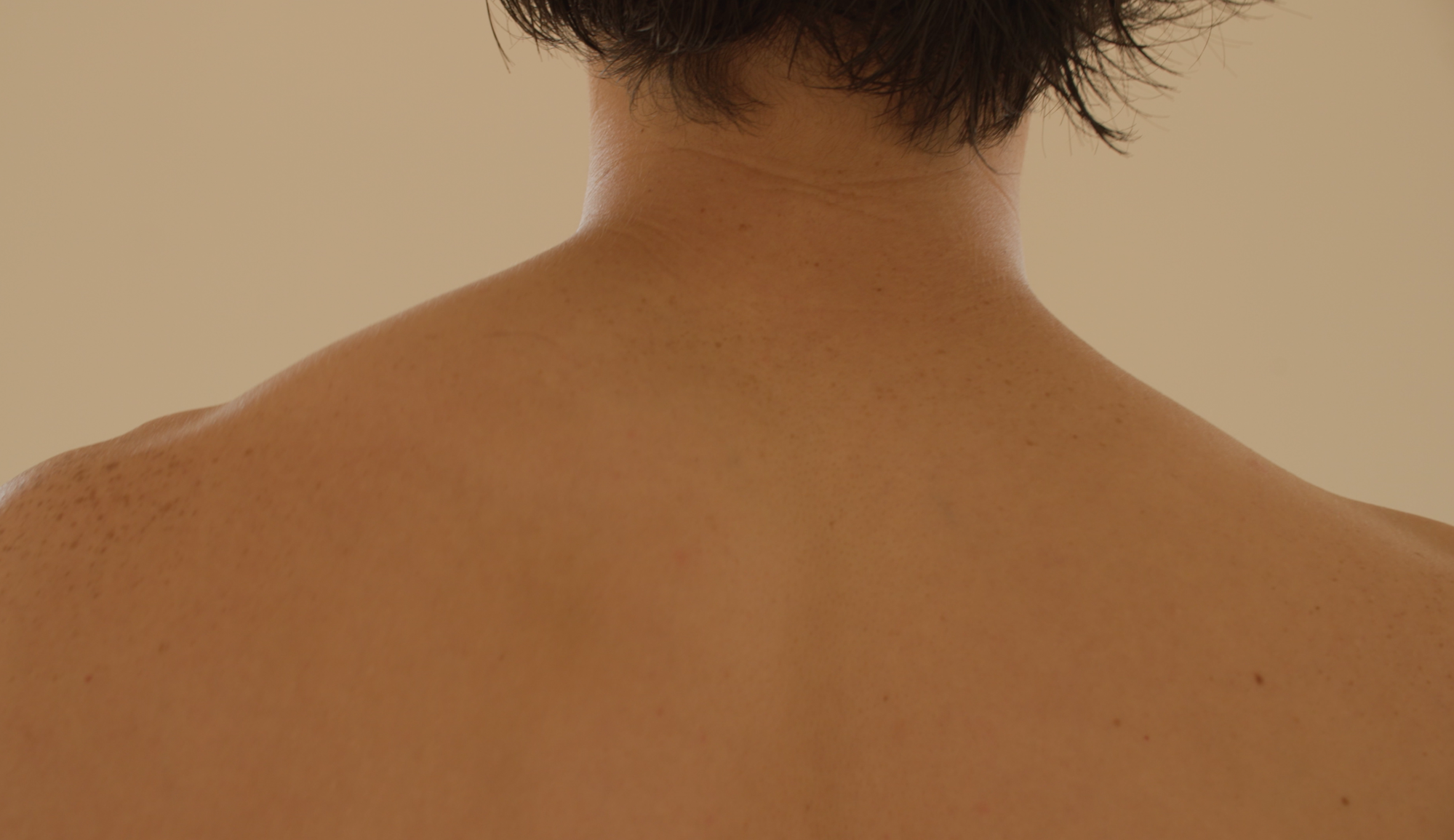 A posterior view of bare shoulders, which span the width of the image. The shoulders are muscular and golden-colored; they are sensual and inviting. A short crop of black hair falls just above the subject's neckline.