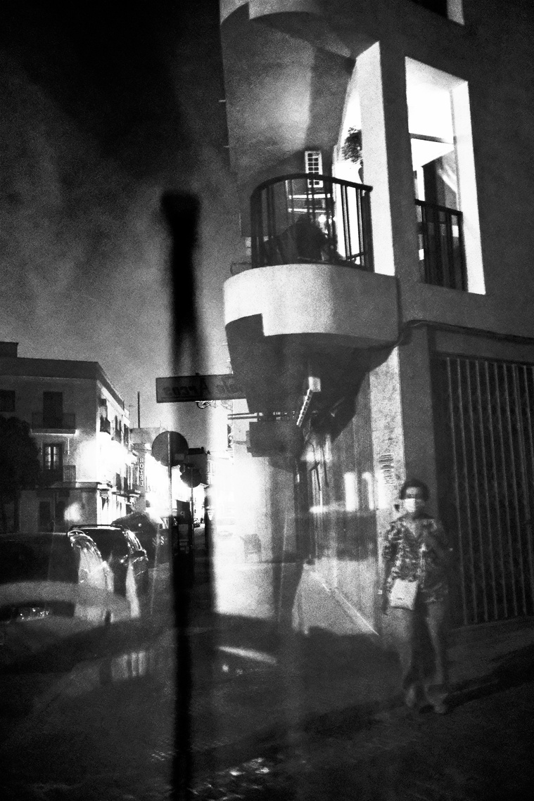 A reflection of a window that appears like curtains.It is a black and white view of a street. You can see Spanish style residences, balconies and cars. A masked woman is walking forwards. Apart from her the streets are empty of people.