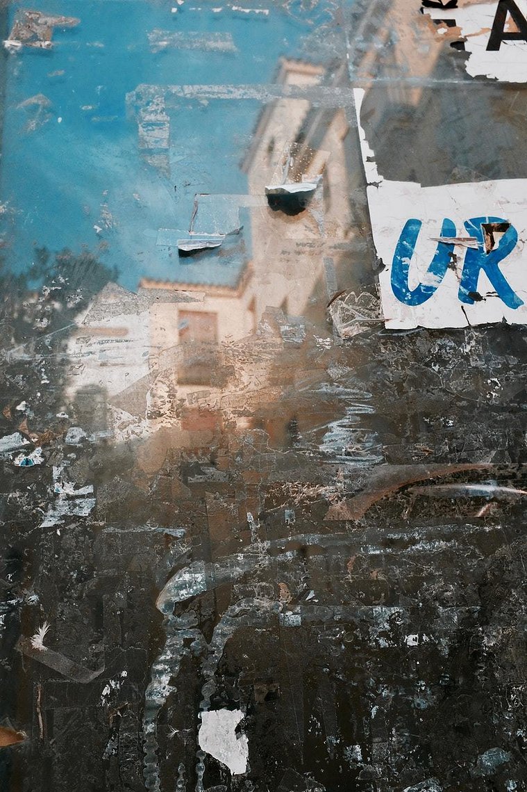 A reflection of houses on mottled metal, with remainders of the marks of glue, paper and stickers on the surface. It looks as if the houses are dissolving. Some walls are white, some are peach coloured, with terracotta roofs and window frames. 