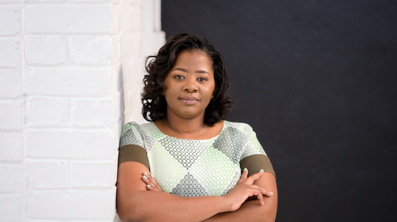 A portrait of Zimbabwe-based photographer, Charmaine Chitate wearing a green checkered dress and leaning against a white wall.