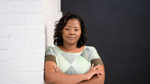 A portrait of Zimbabwe-based photographer, Charmaine Chitate wearing a green checkered dress and leaning against a white wall.