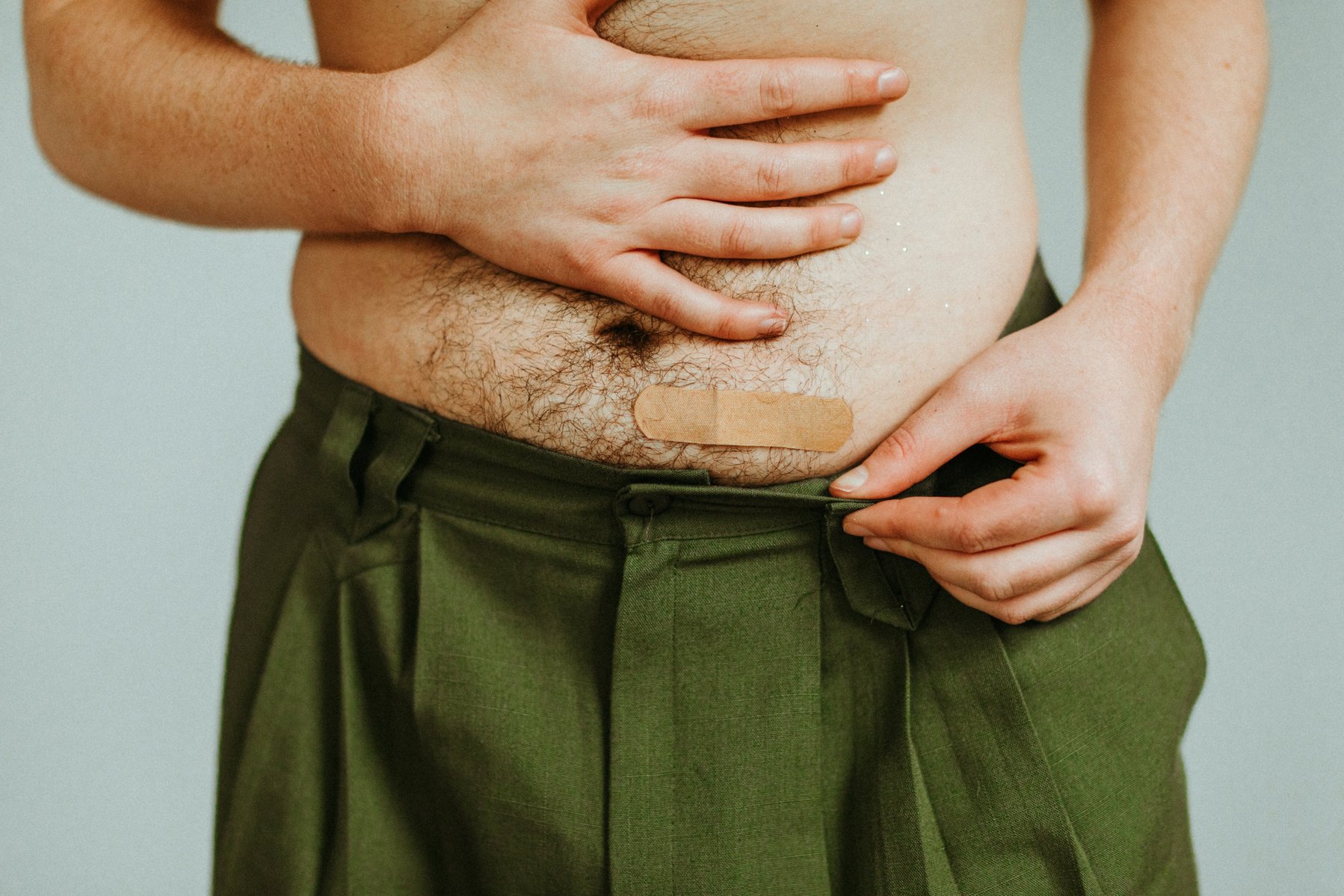 a close-up of Ryan Baker's belly. His belly is pale, covered with dark hair, and has a bandaid covering the lower left side of his belly. The bandaid is covering his injection site. He is wearing earthy green high waisted pants