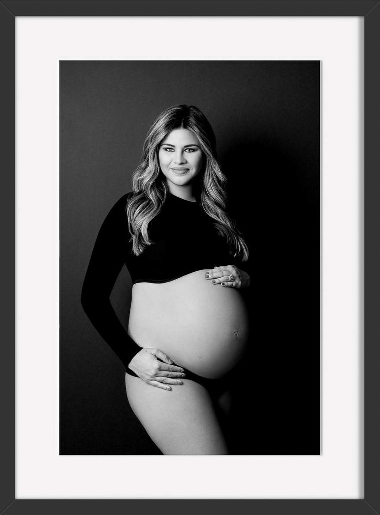 Very pregnant momma to be with long wind blown hair wearing a black long sleeved crop top and black panties with one hand on her pregnant belly and the other under belly and resting on hip with a beautiful smile on her face