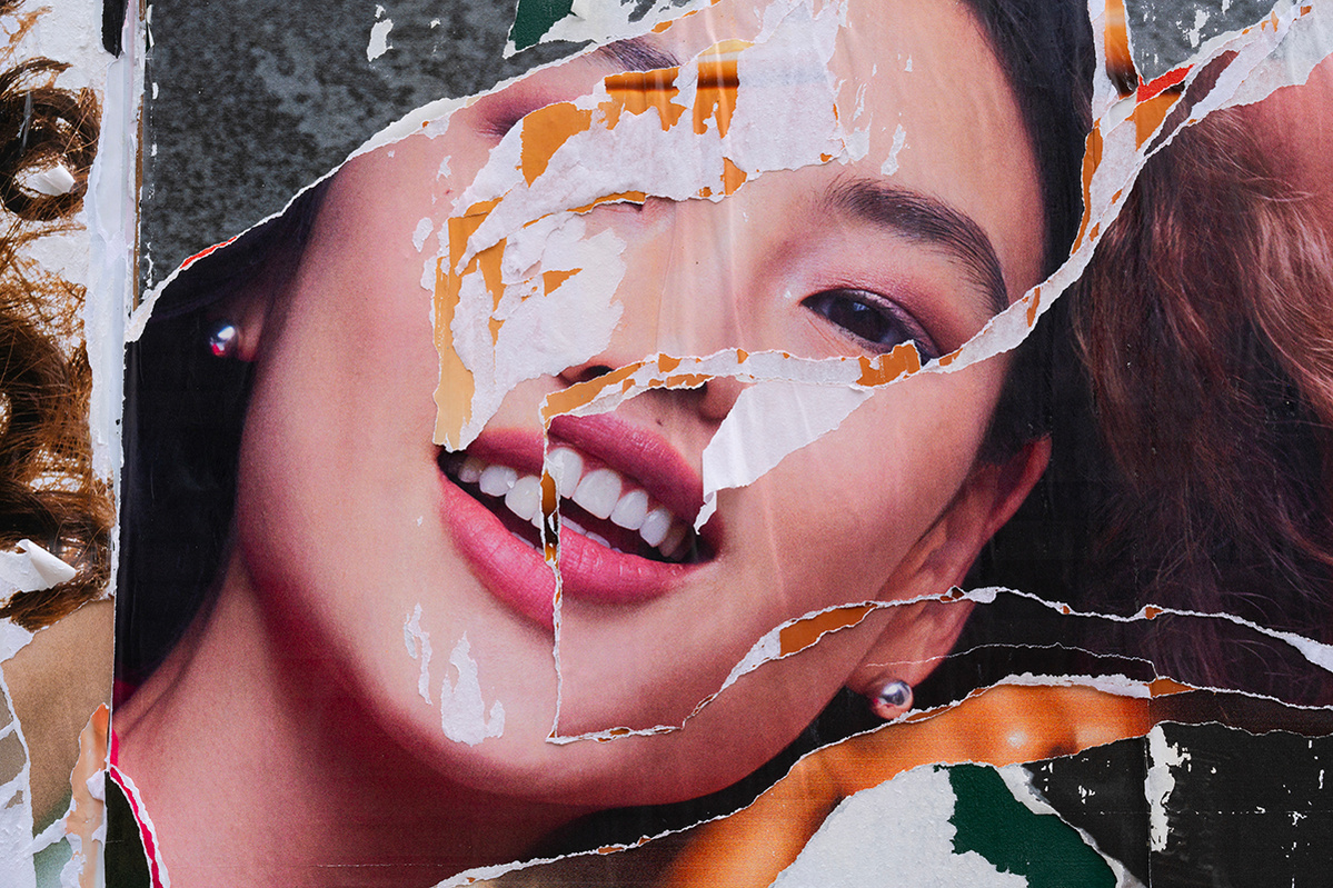 A smiling Asian woman on a shredded advertising poster is seen in double as the first layer of the poster is ripped to reveal an identical one beneath it. One eye is missing, and her face is  not aligned, making the portrait surreal and disconcerting,  