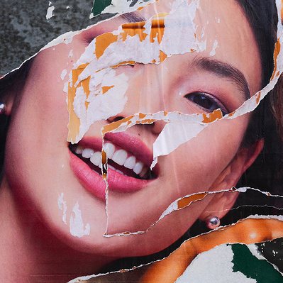 A smiling Asian woman on a shredded advertising poster is seen in double as the first layer of the poster is ripped to reveal an identical one beneath it. One eye is missing, and her face is  not aligned, making the portrait surreal and disconcerting,  