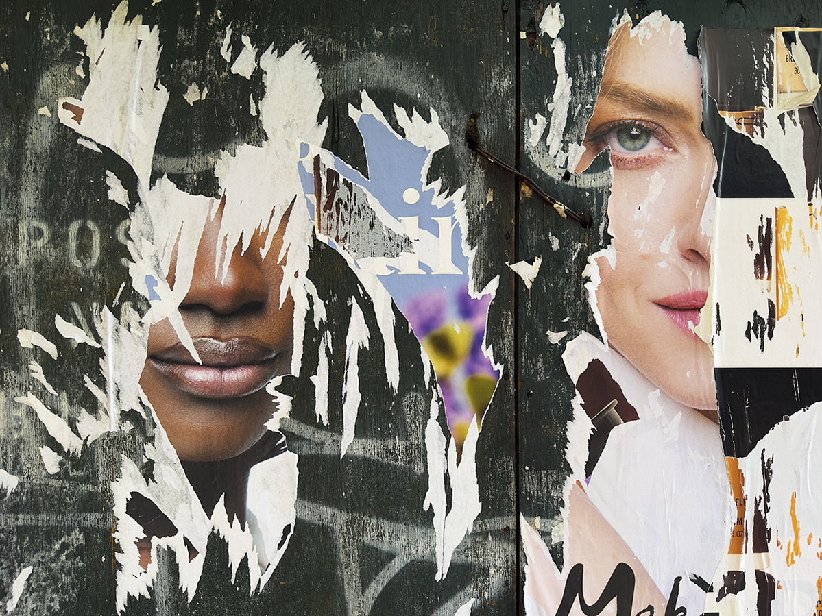 Two mask-like faces emerge from ripped advertising posters on a dark green wooden background. An African-American woman and a White Woman. Only the mouth and nose remain of the black woman. Only one blue eye and part of the lip and nose of the White woman