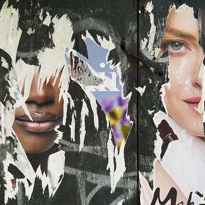 Two mask-like faces emerge from ripped advertising posters on a dark green wooden background. An African-American woman and a White Woman. Only the mouth and nose remain of the black woman. Only one blue eye and part of the lip and nose of the White woman
