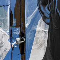 This image features a  segment from a wall of posters and a locked door with a Master lock. The poster is of the side of a Black Man's head, with only one ear showing next to the lock. The highlights on the ear are in blue, reflecting the layered image. 