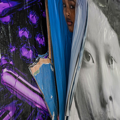 Multiple posters peel off a wall, revealing a black, African-American woman on a color advertisement peering out as if crying, "Help!" The oppressing poster in black and white  [b/w] features a close-up of an Asian woman with a neutral expression.  
