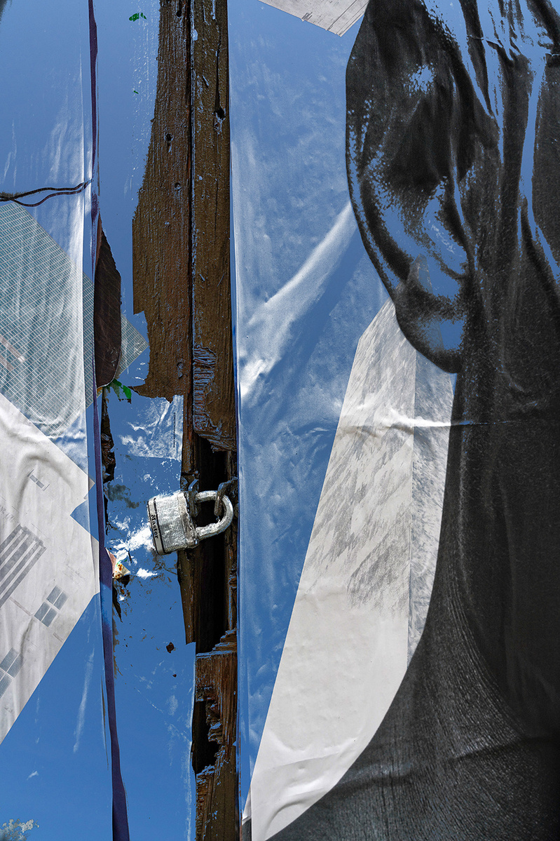 This image features a  segment from a wall of posters and a locked door with a Master lock. The poster is of the side of a Black Man's head, with only one ear showing next to the lock. The highlights on the ear are in blue, reflecting the layered image. 