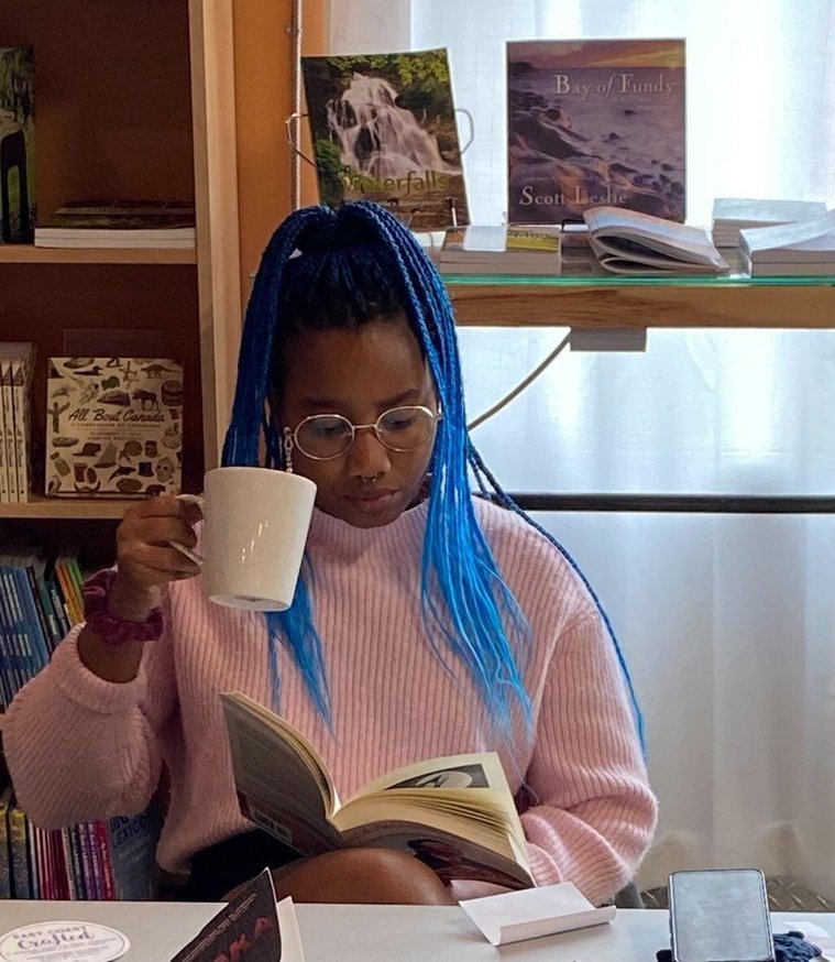 Headshot of Artist: Chloe Bramble holds a white mug in the air by her face while they look down at the book in their other hand. Chloe has long blue twists in a ponytail on top of her head. Behind Chloe is a bookshelf display and a white curtain.