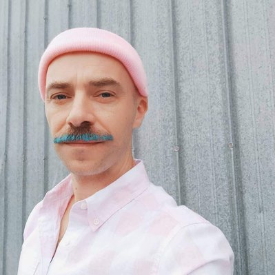 Team Member Headshot: Richie Wilcox looks at the camera with a blue fringed moustache. They are wearing a baby pink toque and a baby pink plaid shirt. The backdrop is a metal ridged wall.