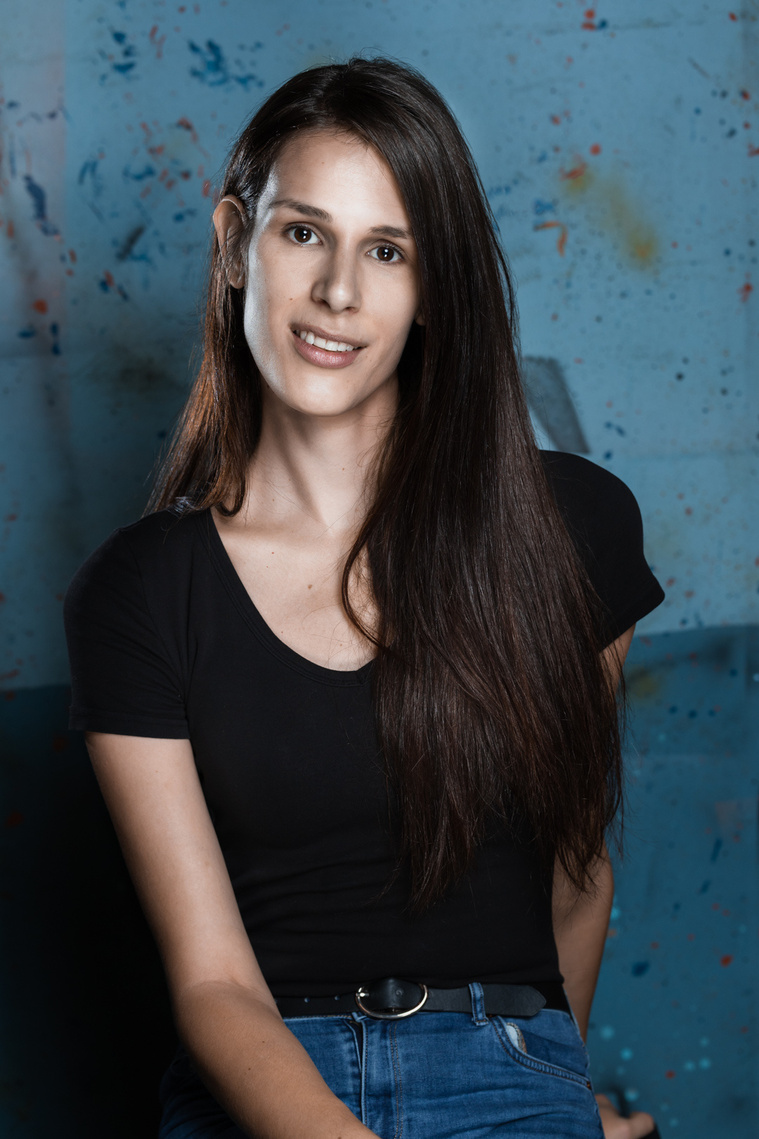 Headshot of Artist: Lara Lewis smiles at the camera. Lara has long brown hair swept to her left shoulder. She is wearing a black t-shirt and blue jeans. The background of the image is a light blue wall with dark blue and orange speckled all over. 