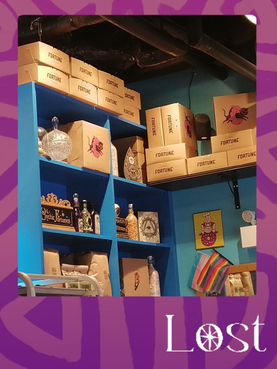 Gallery Image: Text reads: LOST. On a purple border around an image of a blue shelving unit filled with light brown boxes, glass trinkets, clay bottles and card-sized art pieces.