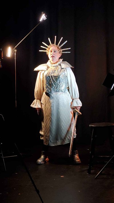 Lou is pictured in their Joan-of-arc attire this time they stand further from the camera and hold their sword down at their side. They are mid-speech. Lou wears healed platform shoes. The backdrop is a black room with two spots of corner lighting.
