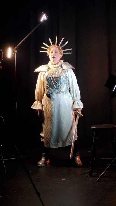 Lou is pictured in their Joan-of-arc attire this time they stand further from the camera and hold their sword down at their side. They are mid-speech. Lou wears healed platform shoes. The backdrop is a black room with two spots of corner lighting.
