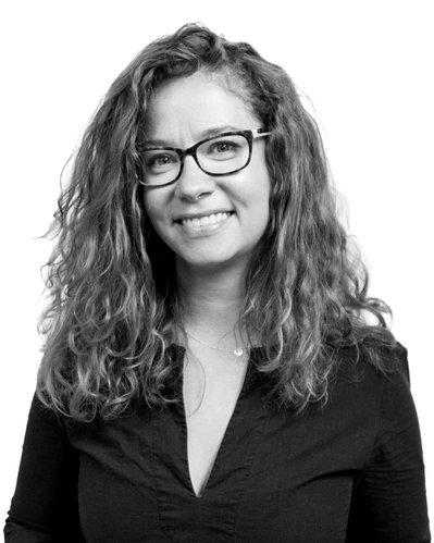 Team Member Headshot: Sylvia bell smiles at the camera. Her curly hair frames her face. Sylvia is wearing curved rectangular glasses and a black blouse. Sylvia's photo floats on a white backdrop.