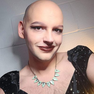 Team Member Headshot: Vie Jones smiles at the camera they wear a sparkled black dress with puffed shoulders and a teal/gold necklace. Vie's head is tilted, their bald head is smooth and they have swooshed eyebrows. The backdrop is a white brick wall.