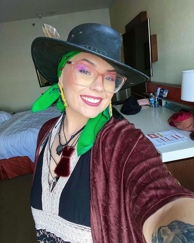 Artist Headshot. Ceilidh Pitaw smiles at the camera wearing a green scarf under a black wide-brimmed hat. Ceilidh takes a selfie with one arm holding the camera in front of her. Ceilidh wears many necklaces including a red pouch. She is in a hotel room.
