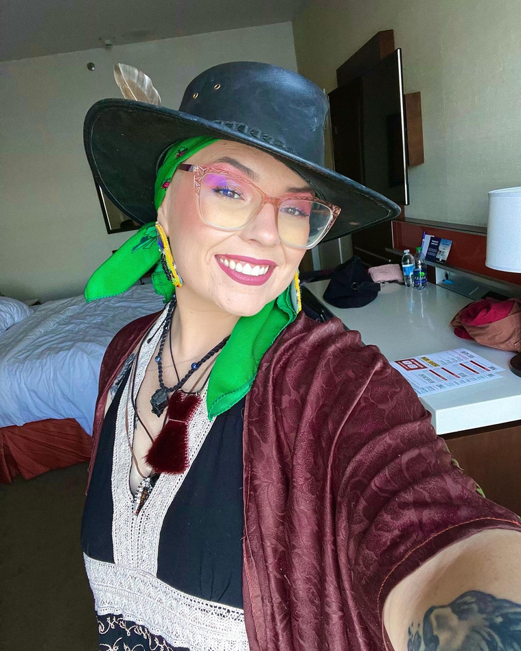 Artist Headshot: Ceilidh Pitaw smiles at the camera wearing a green scarf under a black wide-brimmed hat. Ceilidh takes a selfie with one arm holding the camera in front of them. Ceilidh wears large beaded earrings & many necklaces including a red dress. 