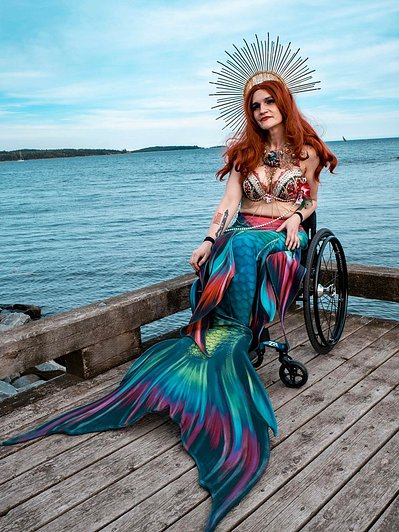 Team Member Headshot: April Hubbard is wearing long red hair, a golden crown, a red and white bejewelled bikini and a multicoloured mermaid tail. They are sitting in their wheelchair on a wooden warf. Behind them is a blue sky and ocean.