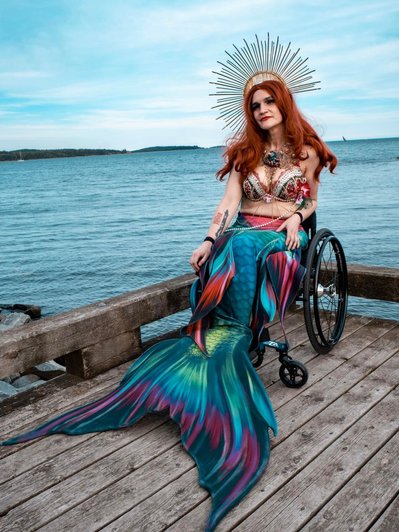 Team Member Headshot: April Hubbard is wearing long red hair, a golden crown, a red and white bejewelled bikini and a multicoloured mermaid tail. They are sitting in their wheelchair on a wooden warf. Behind them is a blue sky and ocean.