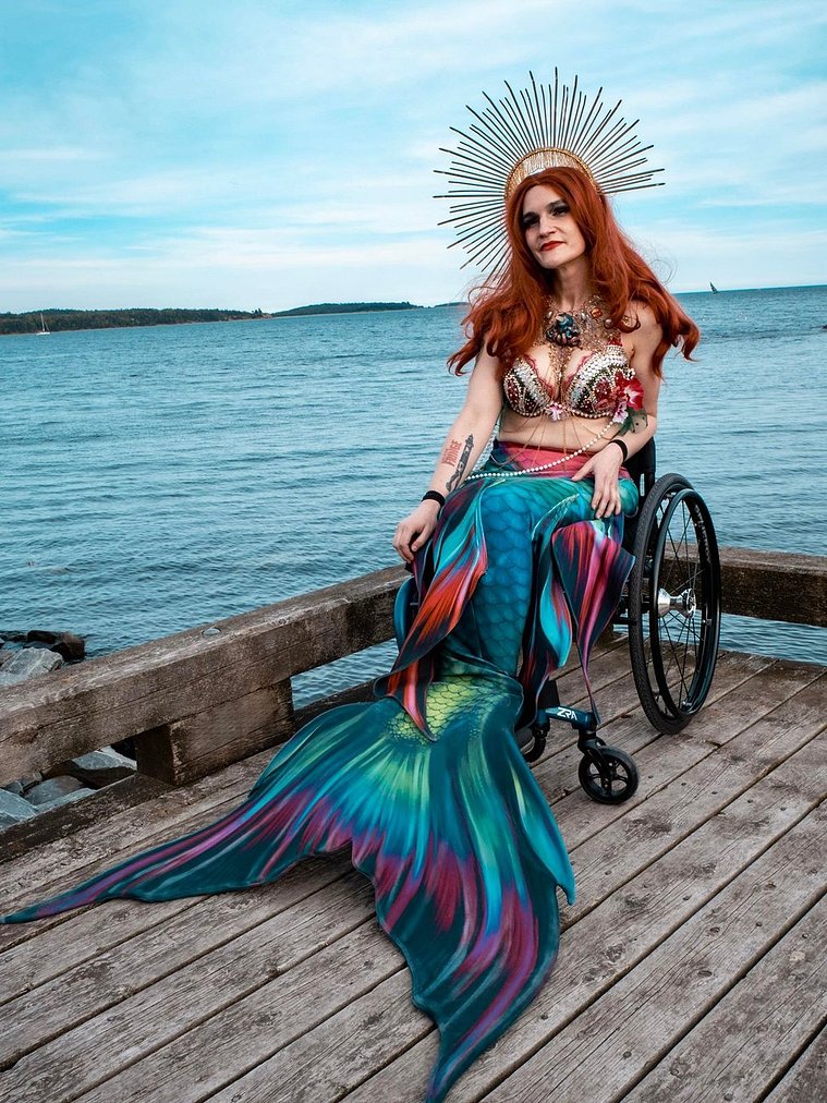 Headshot: April Hubbard is wearing long red hair, a golden crown, a red and white bejewelled bikini and a multicoloured mermaid tail. They are sitting in their wheelchair on a wooden warf. Behind them is a blue sky and ocean.