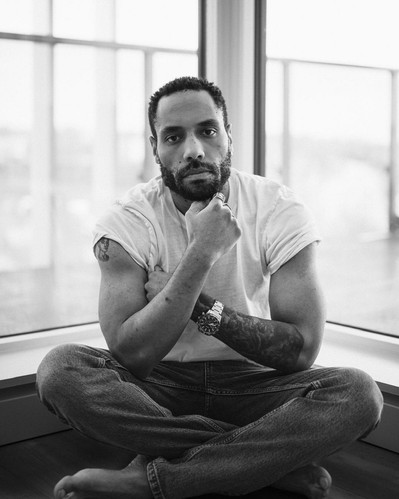 Team Member Headshot: Tyler Simmonds looks at the camera with his hand on his bearded chin. His hair in a coiffured crisp line-up. He wears a white t-shirt with the sleeves pushed up and sits crossed legged in front of a corner window. 