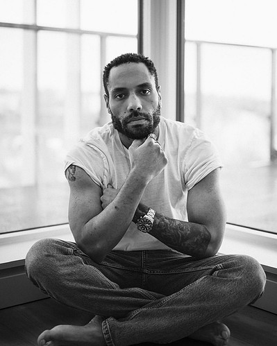 Team Member Headshot: Tyler Simmonds looks at the camera with his hand on his bearded chin. His hair in a coiffured crisp line-up. He wears a white t-shirt with the sleeves pushed up and sits crossed legged in front of a corner window. 